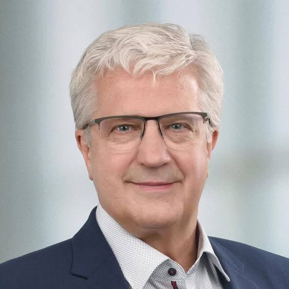 Smiling man with short white hair and glasses, wearing a navy blazer and patterned shirt.